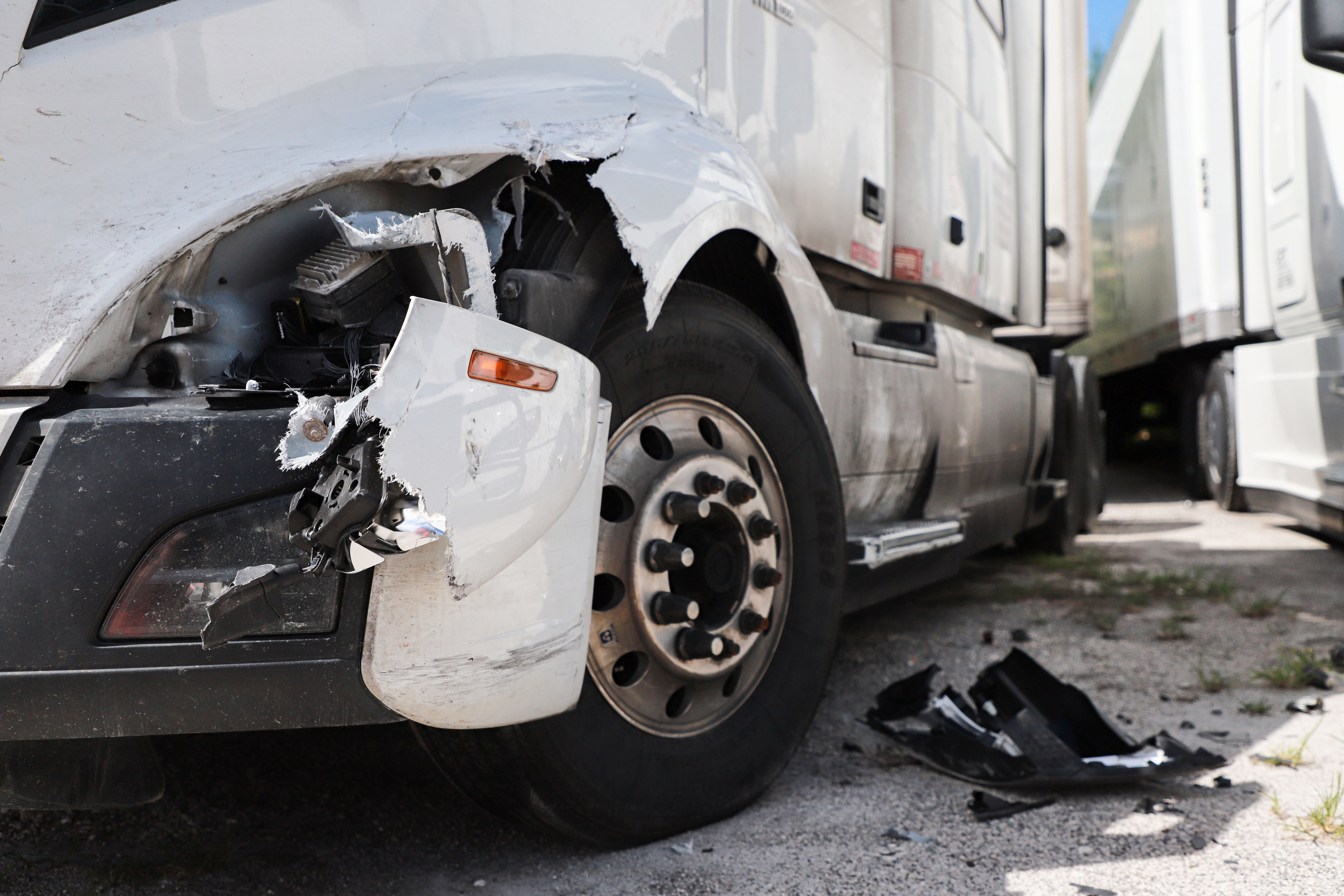 Close-up of damaged front left side of white semi-truck with torn bumper and debris on ground.